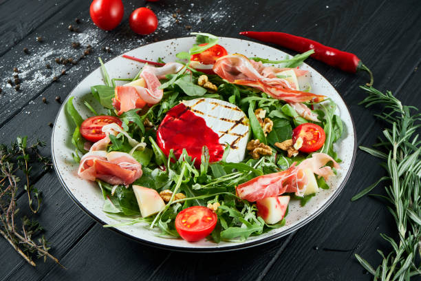 istock Delicious salad with grilled camembert, arugula, spinach, jamon, cherry tomatoes in white on a black background. Close up 1212940543