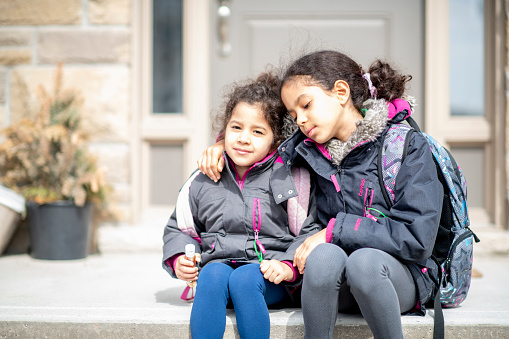 Two Middle Eastern sisters sit on their front porch step together with their backpacks on as they look distraught at the news of school closures during the COVID-19 outbreak.  They are dressed casually and have winter coats on.