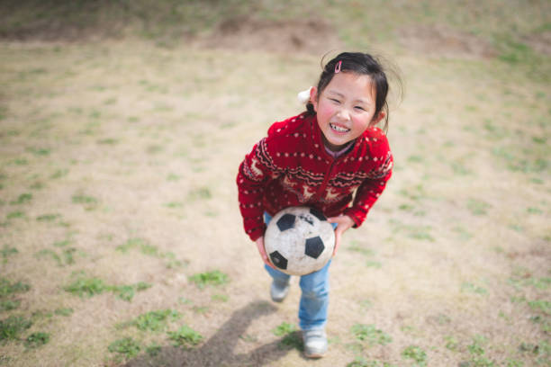 A cheerful child enjoys a playful moment with an adult, who is holding a red soccer ball. The scene takes place outdoors in a vibrant, leafy garden, capturing joy and family bonding.
