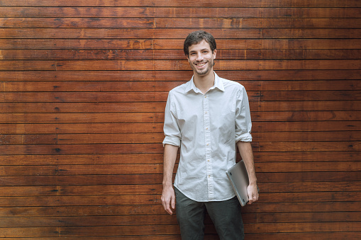 Portrait of creative business man holding laptop while standing against wood paneling in office