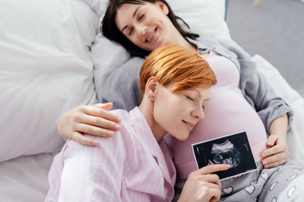 Smiling woman holding ultrasound scan of baby near belly of pregnant girlfriend on bed Smiling woman holding ultrasound scan of baby near belly of pregnant girlfriend on bed surrogacy stock pictures, royalty-free photos & images