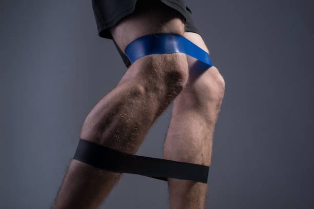 closeup, bottom view of the guys muscular legs in sports shorts, stretching fitness elastic bands with his legs, involvement of leg muscle groups that are not involved in everyday life. On a gray background. Advertising fitness gum. Athletic guy. Pronounced muscles of the legs. primer plano, vista inferior de las piernas musculosas de los chicos en pantalones cortos deportivos, estirando bandas elásticas de fitness con sus piernas, implicación de grupos musculares de piernas que no están involucrados en la vida cotidiana.