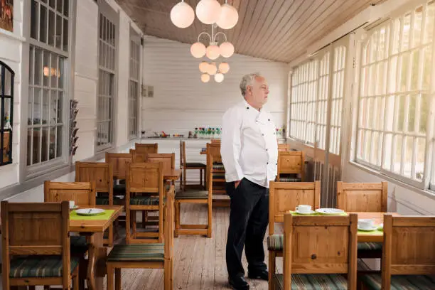 A concept to illustrate the economic impact of the Covid-19 virus on the restaurant and catering business. Restaurant owner wearing his chef’s whites standing in his empty restaurant. Photographed on location in a restaurant on the island of Møn in Denmark.