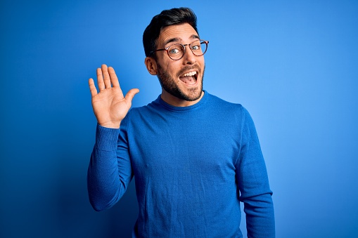 Joven guapo hombre con barba usando suéter casual y gafas sobre el fondo azul Waiving diciendo hola feliz y sonriente, gesto de bienvenida amistoso photo