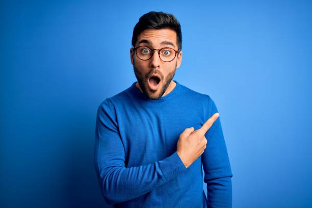 Photo of Young handsome man with beard wearing casual sweater and glasses over blue background Surprised pointing with finger to the side, open mouth amazed expression.