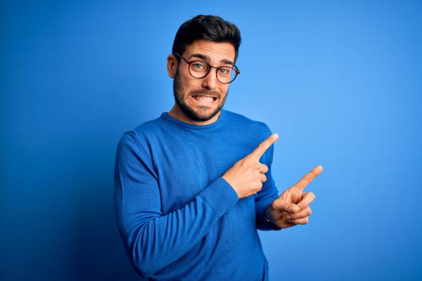 Young handsome man with beard wearing casual sweater and glasses over blue background Pointing aside worried and nervous with both hands, concerned and surprised expression Young handsome man with beard wearing casual sweater and glasses over blue background Pointing aside worried and nervous with both hands, concerned and surprised expression bad teeth stock pictures, royalty-free photos & images