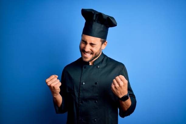 Young handsome chef man with beard wearing cooker uniform and hat over blue background excited for success with arms raised and eyes closed celebrating victory smiling. Winner concept.
