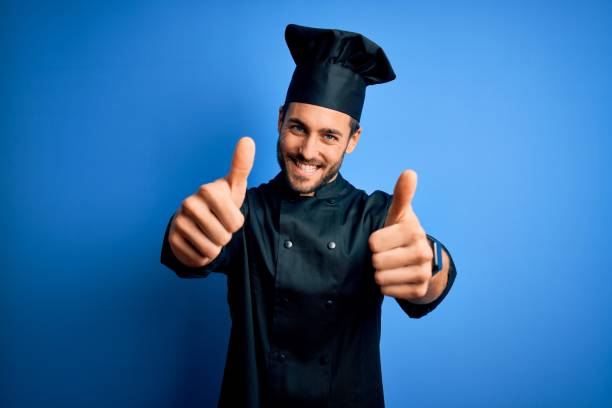 istock Young handsome chef man with beard wearing cooker uniform and hat over blue background approving doing positive gesture with hand, thumbs up smiling and happy for success. Winner gesture. 1212961131