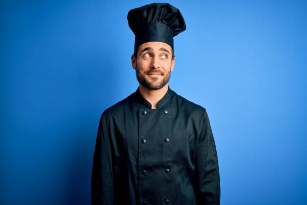 young handsome chef man with beard wearing cooker uniform and hat over blue background smiling looking to the side and staring away thinking. - mannered imagens e fotografias de stock