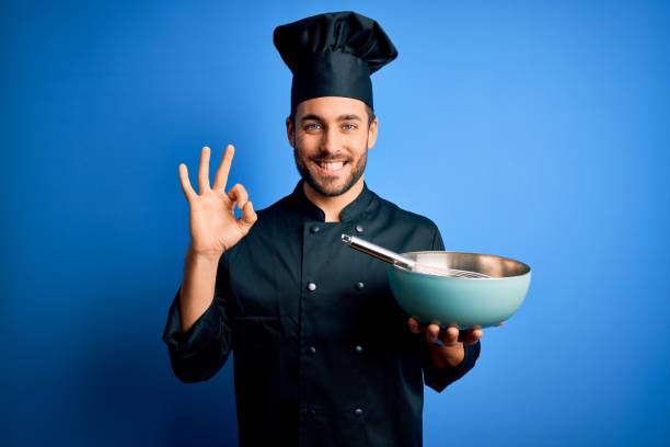 Young cooker man with beard wearing uniform using whisk and bowl over blue background doing ok sign with fingers, excellent symbol Young cooker man with beard wearing uniform using whisk and bowl over blue background doing ok sign with fingers, excellent symbol ok sign photos stock pictures, royalty-free photos & images