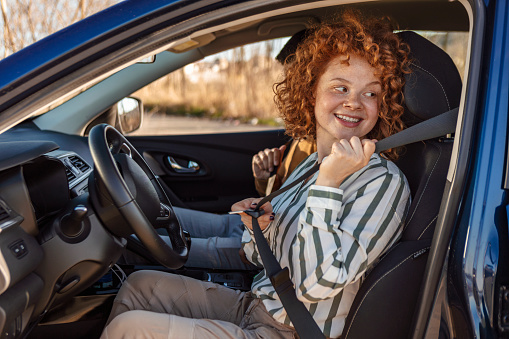 Car driver young woman wearing safety belt driving new blue car in summer. Happy redhead female looking back.