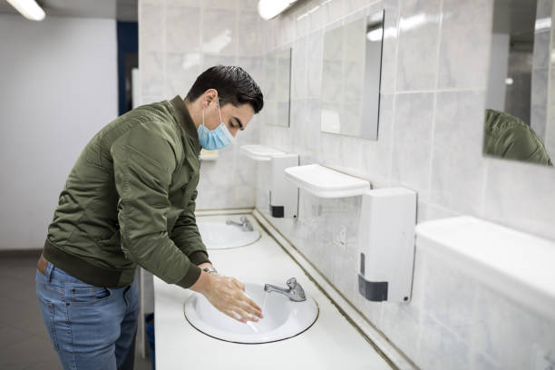 Man with a face mask washes his hands in a public bathroom