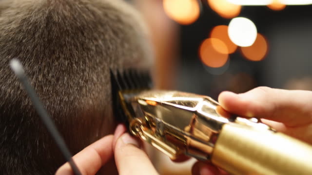 Trendy barber cuts bearded man's hair with a clipper in barbershop. Men's hairstyling and hair cutting in salon. Grooming the hair with trimmer. Hairdresser doing haircut in retro hair salon