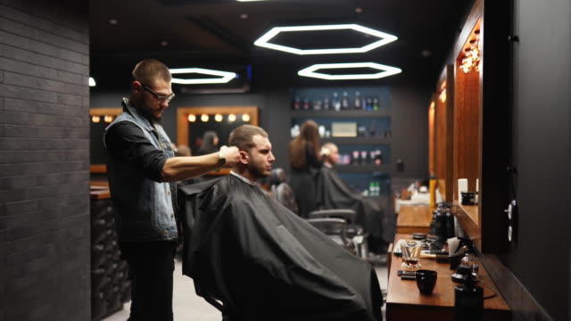 Trendy barber cuts bearded man's hair with a clipper in barbershop. Men's hairstyling and hair cutting in salon. Grooming the hair with trimmer. Hairdresser doing haircut in retro hair salon. Tracking