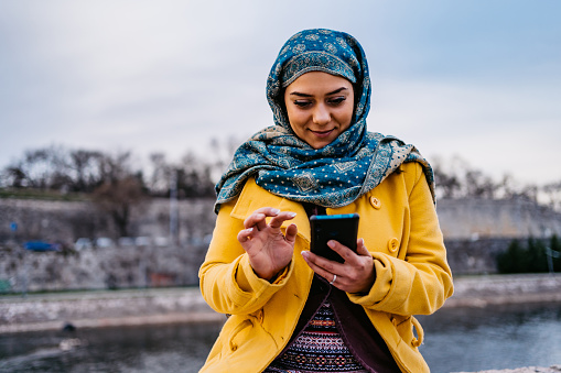Young beautiful smiling Muslim woman with headscarf using smartphone and sitting on quay.