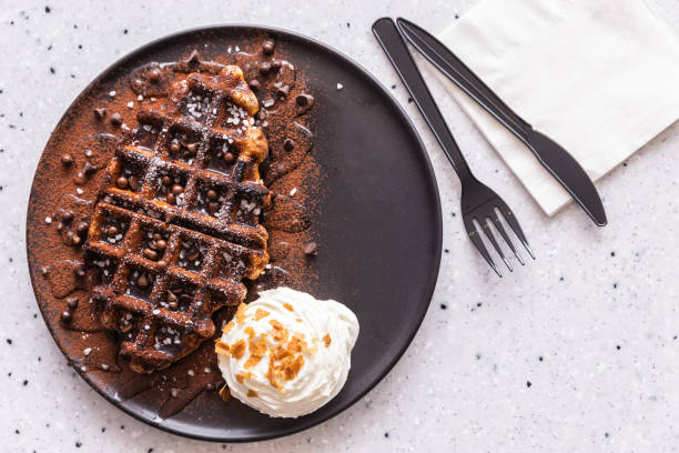 flat lay of yummy homemade belgian waffle topping with dark chocolate syrup, cocoa powder and chocolate chips, served with whipped cream in brown color plate. delicacy and stylish cuisine. - plate changing imagens e fotografias de stock