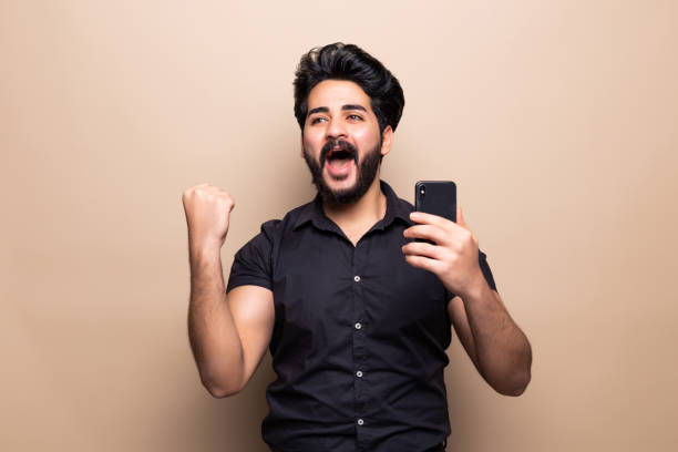 istock Happy bearded young man with victory gesture holding phone isolated on beige background 1212992824