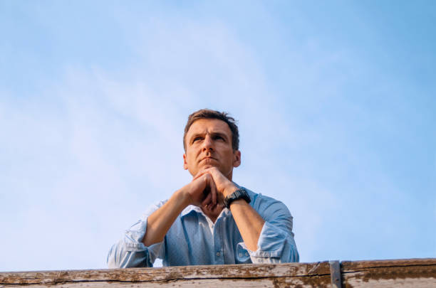 Outdoor portrait of thinking and looking away caucasian adult man wearing white t-shirt against plant wall of green leaves. Eco concept. Sustainable lifestyle.