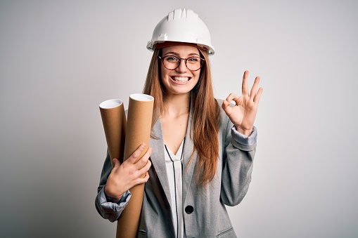 Joven hermosa mujer arquitecta pelirroja que lleva gafas y casco de seguridad sosteniendo planos haciendo bien signo con los dedos, excelente símbolo photo
