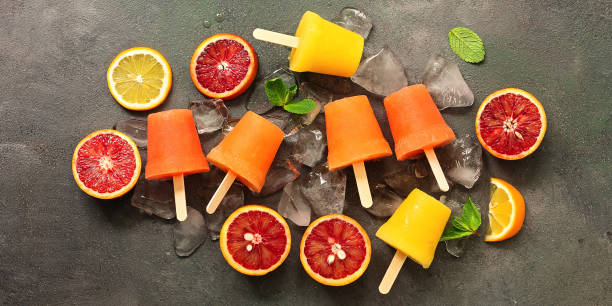 istock Assorted orange popsicles with slices of citrus, mint and ice on a dark rustic background, border. Frozen juice of red and yellow orange on a stick. Top view, flat lay. 1213000329