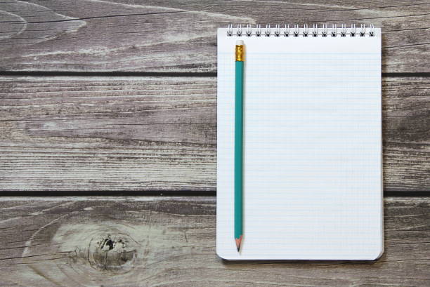 istock Notepad with a blank white sheet in a checker paper with simple graphite pencil lies on the background of wooden boards 1213000577