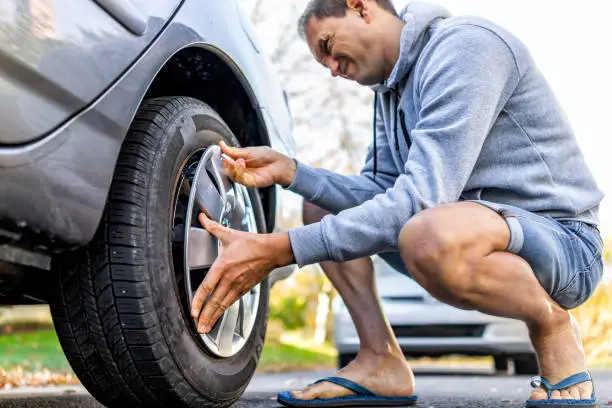 Missing cap cover on parked car tire wheel with man replacing hubcap with new shiny metal outside with difficulty