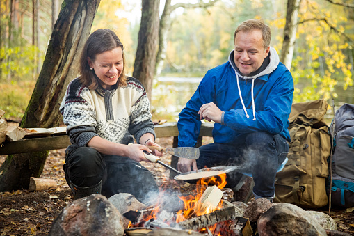 Man and woman making pancakes on campfire in forest on shore of lake, making a fire, grilling. Happy couple exploring Finland. Scandinavian landscape.