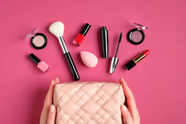 Female hands holding makeup bag with cosmetic products on pink background. Flat lay, top view. Beauty and fashion concept.