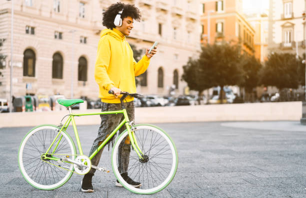happy afro man using mobile smartphone outdoor - young guy having fun listening music with headphones while riding with bike in city - youth millennial generation lifestyle and technology concept - cycling bicycle hipster urban scene imagens e fotografias de stock