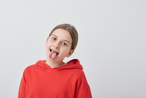 Funny teenage girl in red sweatshirt showing tongue while grimacing in front of camera against white background in isolation