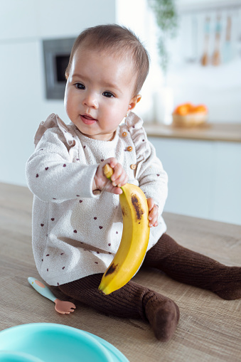 Shot of a cute little girl looking to the side and holding a banana while sitting on the kitchen counter at home.