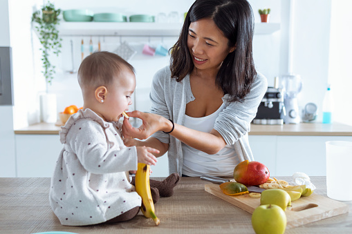 Shot of happy young mother feeding her cute baby girl with a banana in the kitchen at home.