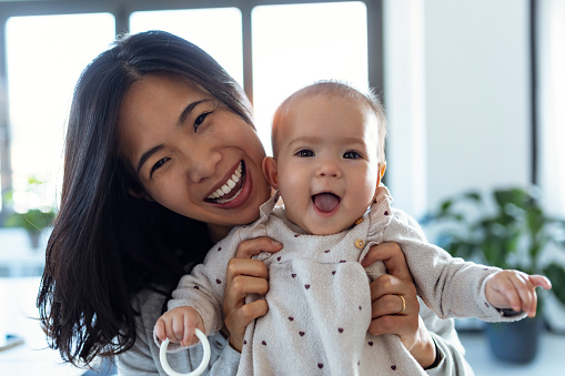 Shot of happy young mother with her baby daughter looking at camera while staying at home.