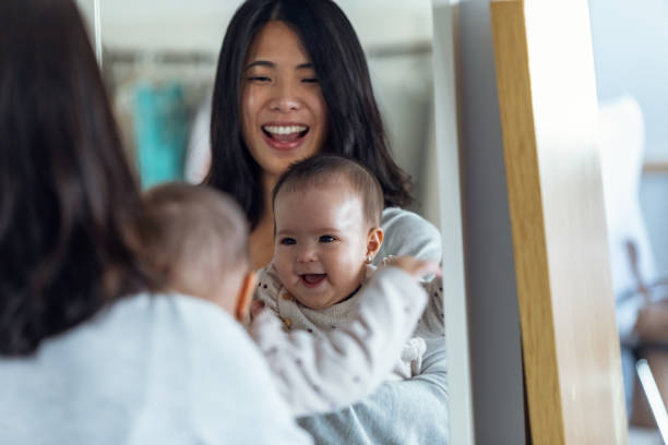 glückliche junge mutter mit ihrem kleinen mädchen, das in den spiegel schaut, während sie zu hause lächelt. - child asian ethnicity little girls toddler stock-fotos und bilder