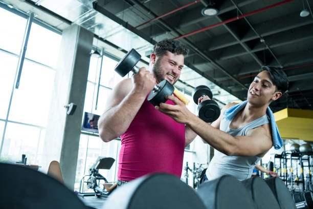 istock Fat man lifting weights with the help of his trainer 1213035547