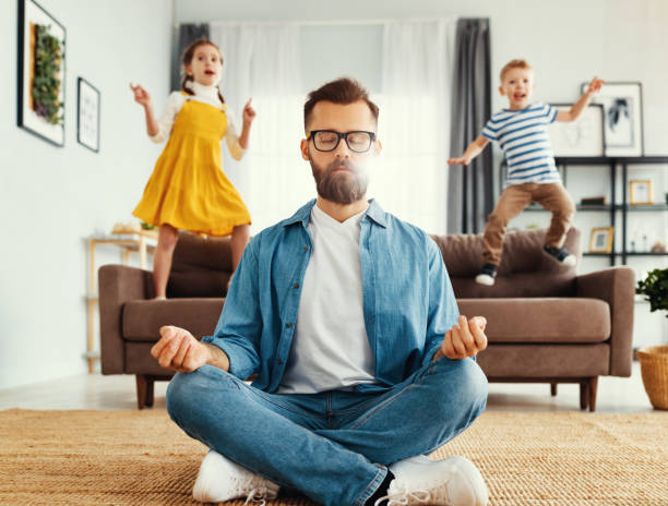 Father meditating in room with playful kids Tranquil young man in casual wear sitting on floor in lotus position and meditating while kids having fun and jumping on sofa at home teasing photos stock pictures, royalty-free photos & images