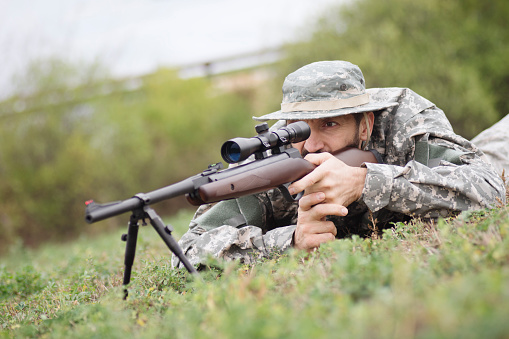 The hunter or soldier in military uniform is aiming and shooting with sniper and shotgun outdoors on grass.