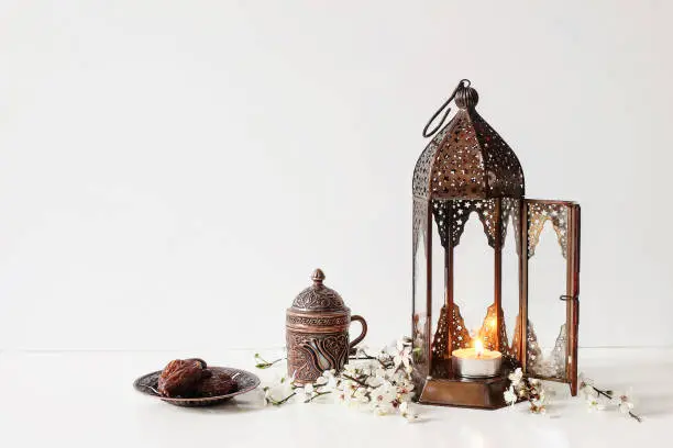 Photo of Ramadan Kareem greeting card, invitation. Bronze plate with dates fruit, white flowers, prunus tree blossoms, cup of tea and glowing Moroccan lantern on table background. Iftar dinner. Eid ul Fitr.