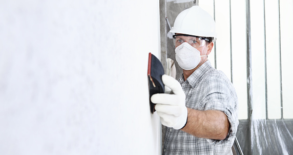 man sand the wall with sandpaper, professional construction worker with mask, safety hard hat, gloves and protective glasses. interior building site, copy space background
