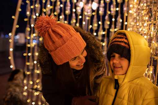 A mom is smiling at her son who is admiring the Christmas lights at night