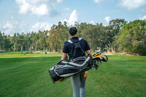 Rear view of a young golfer walking by carrying golf bag in a golf course. Horizontal shot.