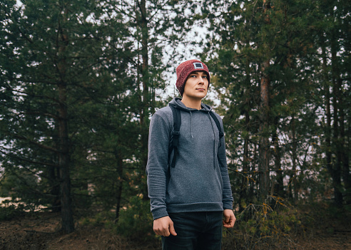 Man in hoodie and a red hat with a backpack is standing on pine forest background