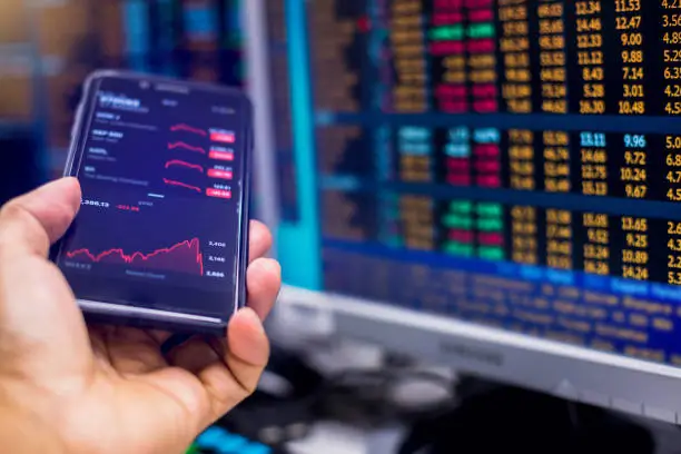 Photo of Red graph on smartphone with blurry computer monitor showing stock price slump.