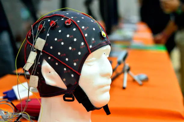Electroencephalogram (EEG) head cap with flat metal discs (electrodes) attached to a white head model. Laptops blurred at background.