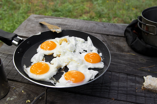 Fried eggs cooked in frying pan on camping gas stove on old wooden table. Outdoor cooking at forest.