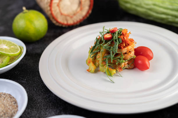 Spicy lime pork salad with bitter melon, galangal, chili, tomatoes and garlic in a white plate on a black cement floor.