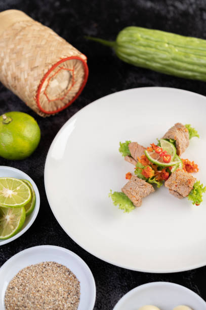 Spicy lime pork salad with bitter melon, galangal, chili, tomatoes and garlic in a white plate on a black cement floor.