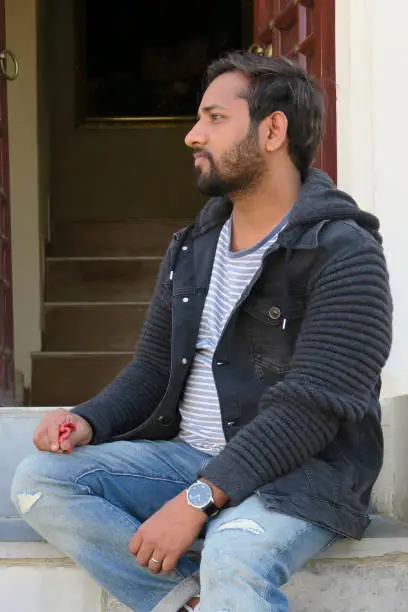 Photo of Image of young handsome Indian man in early 20s posing sitting on staircase looking sideways, thinking / pensive at holiday having fun, good looking man with beard messing about on holiday vacation, wearing black hoodie, ripped jeans, blue white T-shirt