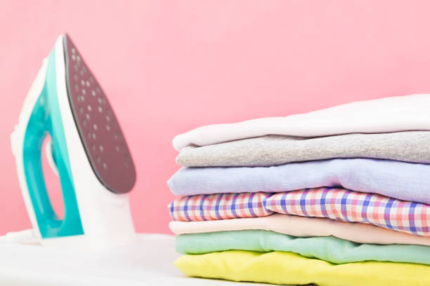 istock Modern Electric iron and stack of clothes on ironing board on pink background in laundry room. Household laundry ironing Woman holding Stacks of folded washed and dried clothes pile 1213125554