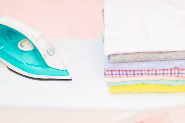 istock Modern Electric iron and stack of clothes on ironing board on pink background in laundry room. Household laundry ironing Woman holding Stacks of folded washed and dried clothes pile 1213125571
